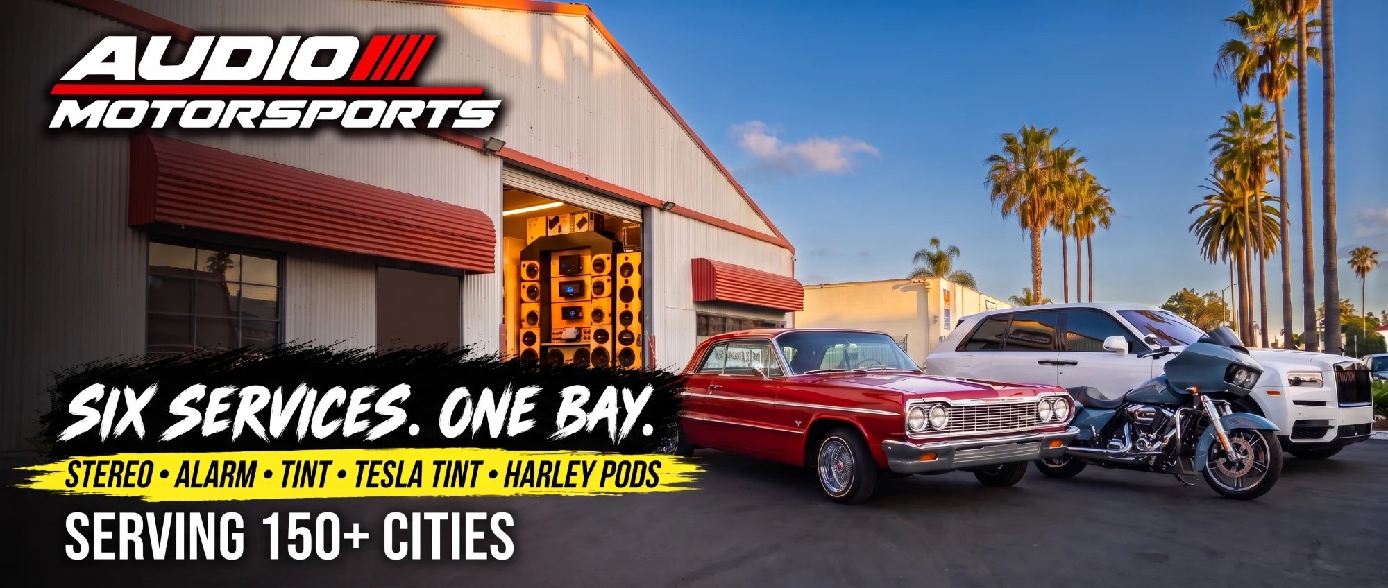 El taller de Audio MotorSports en Carson al atardecer: la bahía abierta con la pared de bocinas, un Impala rojo, una Harley bagger y una SUV blanca al frente
